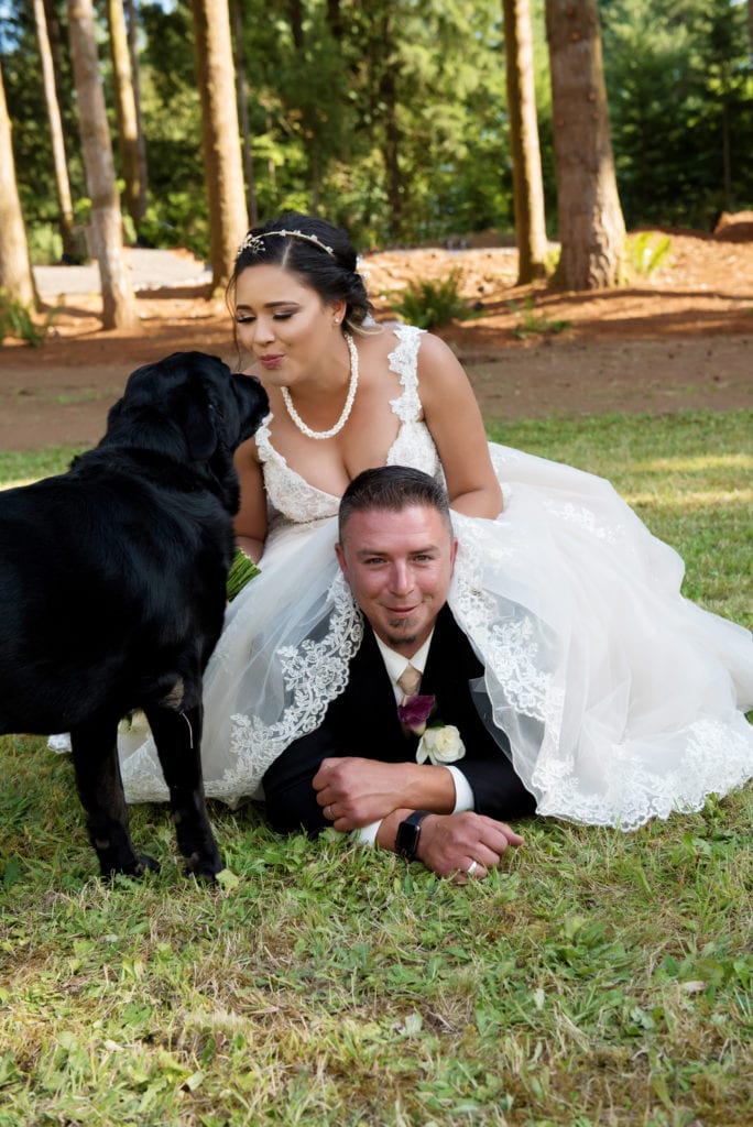 the bride sits on top of the grooms back and kisses their black lab dog
