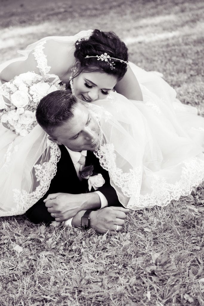 a groom lays in the grass underneath the bride's dress while she leans down around his face