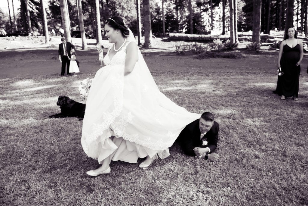a bride in a large wedding dress walks over the groom as he lays on his stomach in the grass
