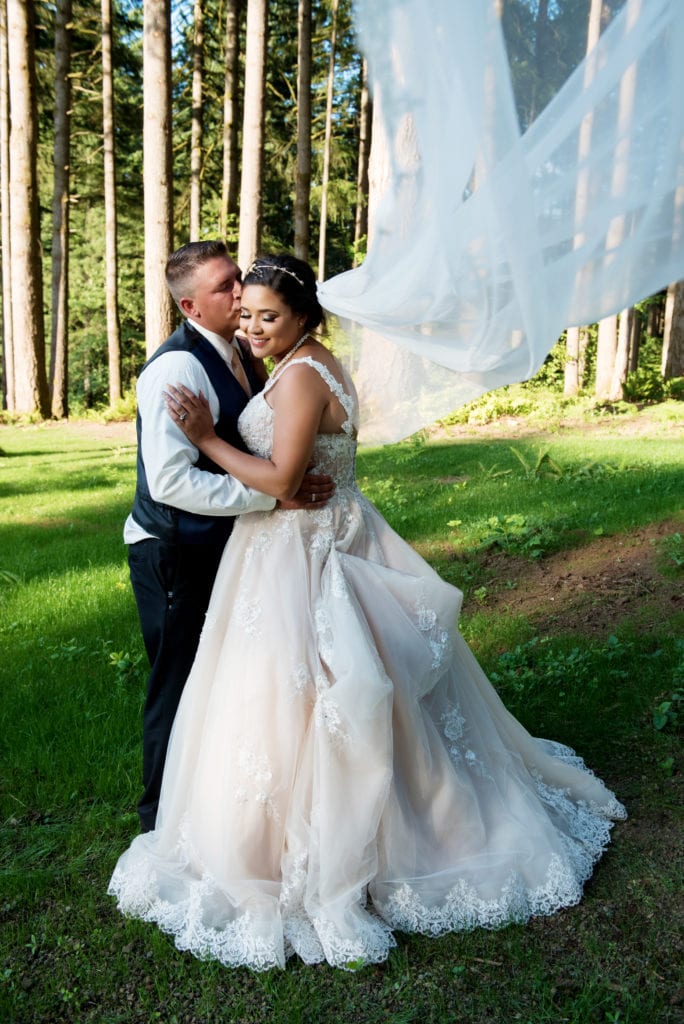 bride and groom kiss in front of tall trees after their brush prairie backyard wedding and the bride's very long veil flies in the wind