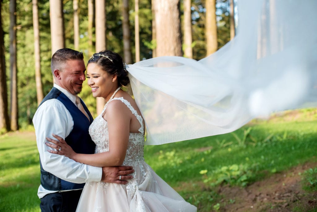 bride and groom laugh in front of tall trees after their brush prairie backyard wedding and the bride's very long veil flies in the wind