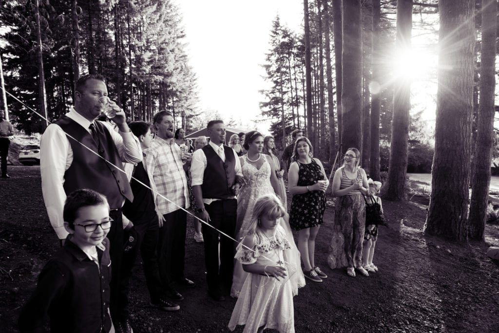 the bride and groom and wedding guests gather around to watch a surprise as the sun shines brightly through the trees
