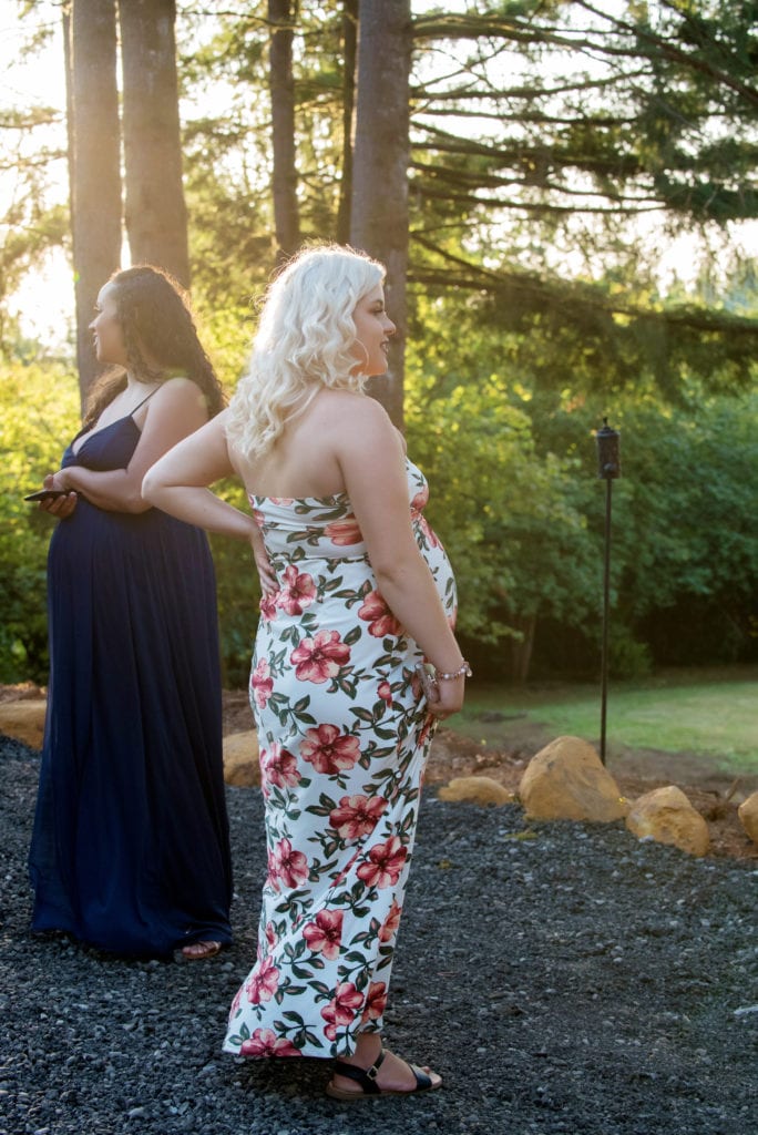 a candid photo of two pregnant women at a wedding standing almost back to back looking opposite directions wearing long dresses
