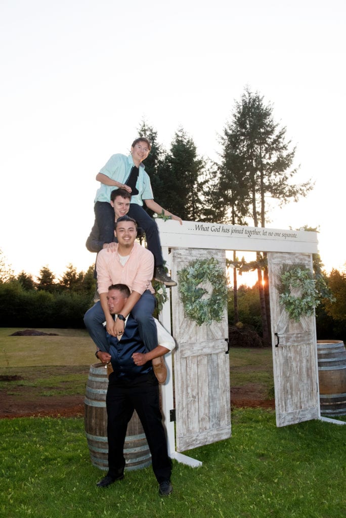 a groom's 3 adult sons climb onto his shoulders after a wedding next to an arbor made of doors