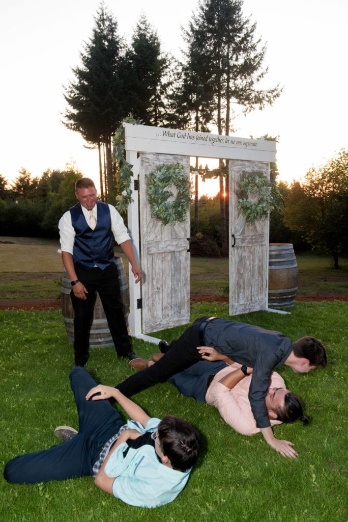 a groom's 3 adult sons climb onto his shoulders and then they have all fallen onto the grass